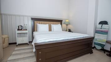 Una cama bien hecha con sábanas blancas, un cabecero de madera, una mesita de noche con una lámpara y un pequeño estante con una tetera y botellas.