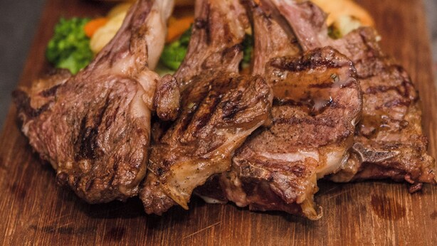 Tagliere di legno con costolette d'agnello alla griglia e un cucchiaio di salsa che vi viene versata sopra.