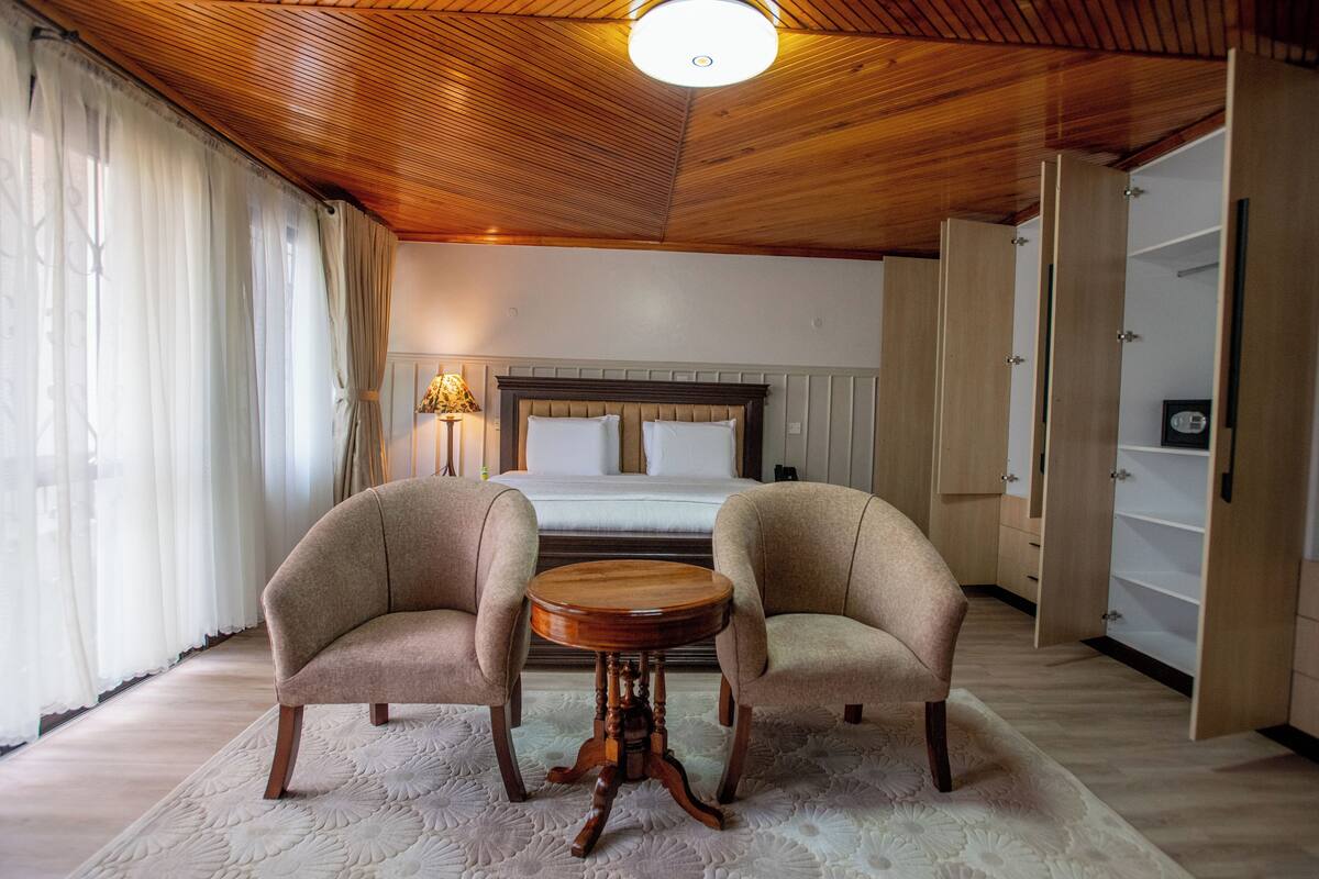 Ein Hotelzimmer mit Holzboden, einem Bett mit weißen Leinen, zwei Sesseln, einem runden Holztisch und einem großen Kleiderschrank.