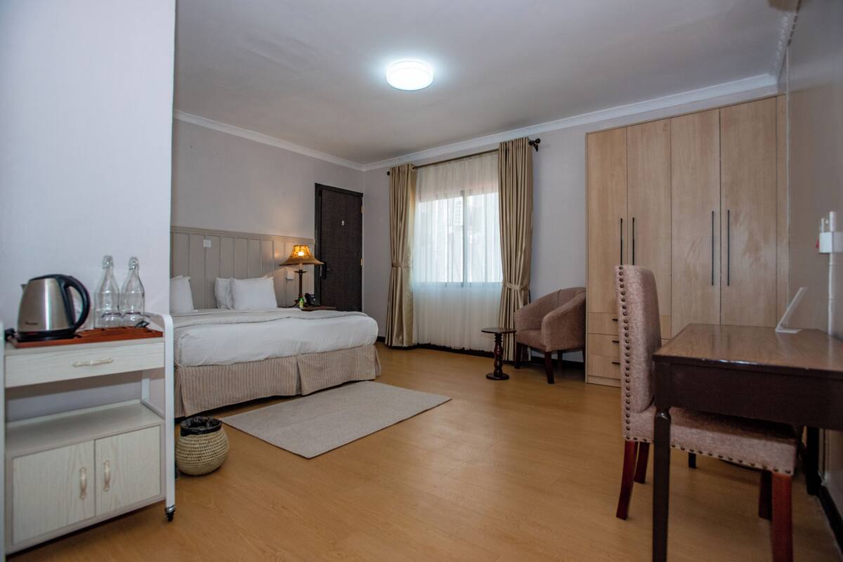 Ein Hotelzimmer mit Bett, Nachttisch, Lampe, Sessel, Schreibtisch, Kleiderschrank und einem fenster mit Vorhängen.