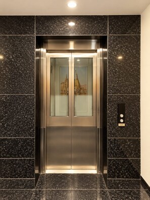 An elevator with a metallic finish, a glass panel with a cityscape view, and a control panel with buttons. - Laxmi Vilas (Varanasi)