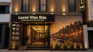 A hotel named Laxmi Vilas Stay with a modern facade, multiple windows, and a sign indicating amenities like AC rooms, free Wi-Fi, 24x7 service, and clean & safe environment. - Laxmi Vilas (Varanasi)