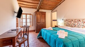 Una habitación con techo de madera, cama con colcha azul turquesa, armario de madera, escritorio con silla y televisor montado en la pared.