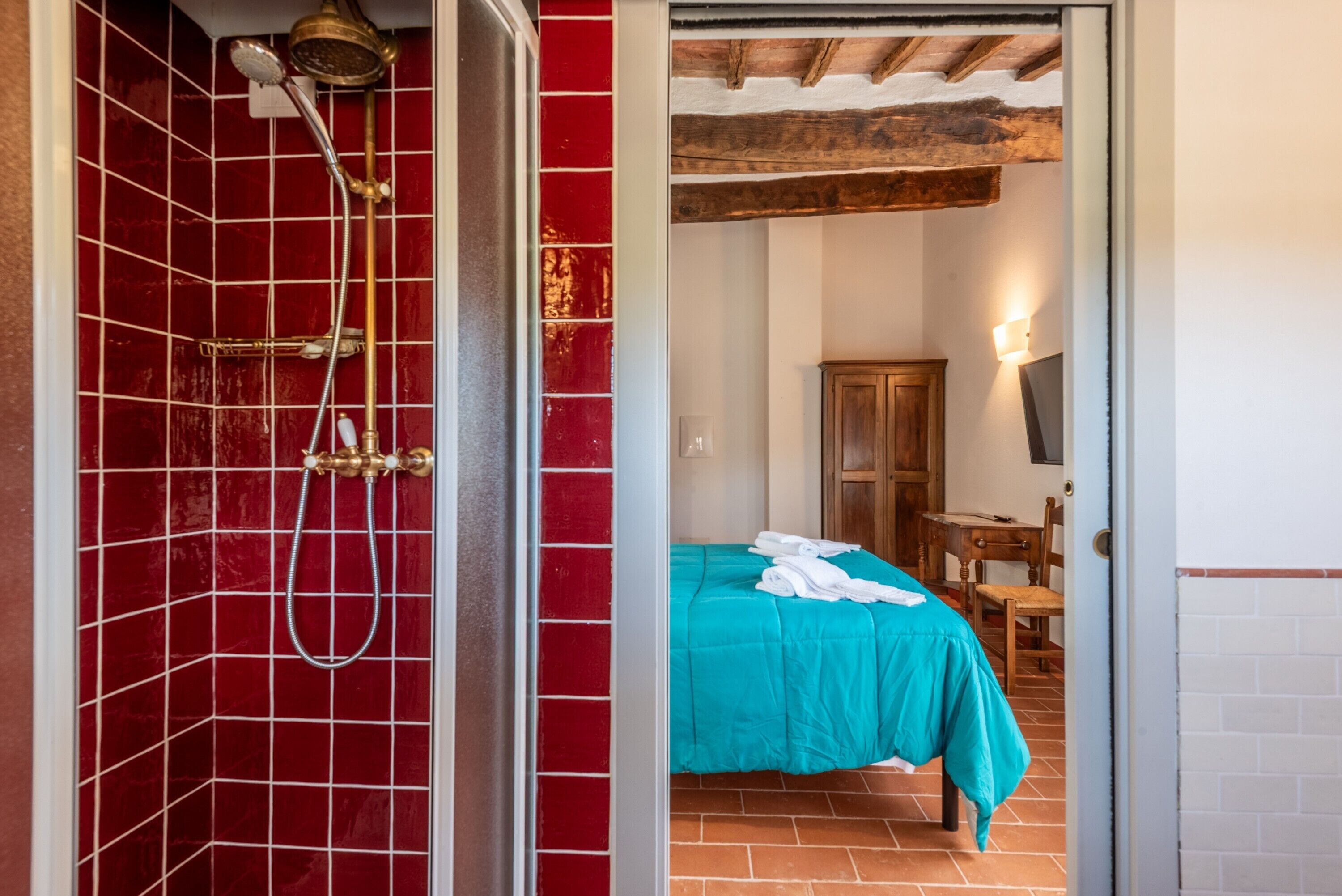 Una habitación con ducha de azulejos rojos, una cama con colcha color turquesa, un armario de madera y una mesa con una silla.