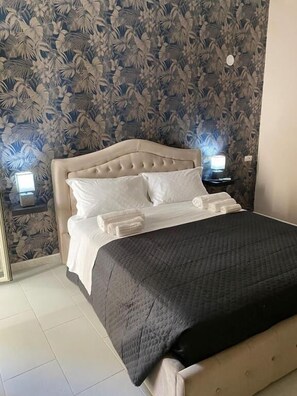 A neatly made bed with a dark quilt, white pillows, and a beige headboard. There are two bedside lamps on either side of the bed. - Melody rooms (Syracuse)