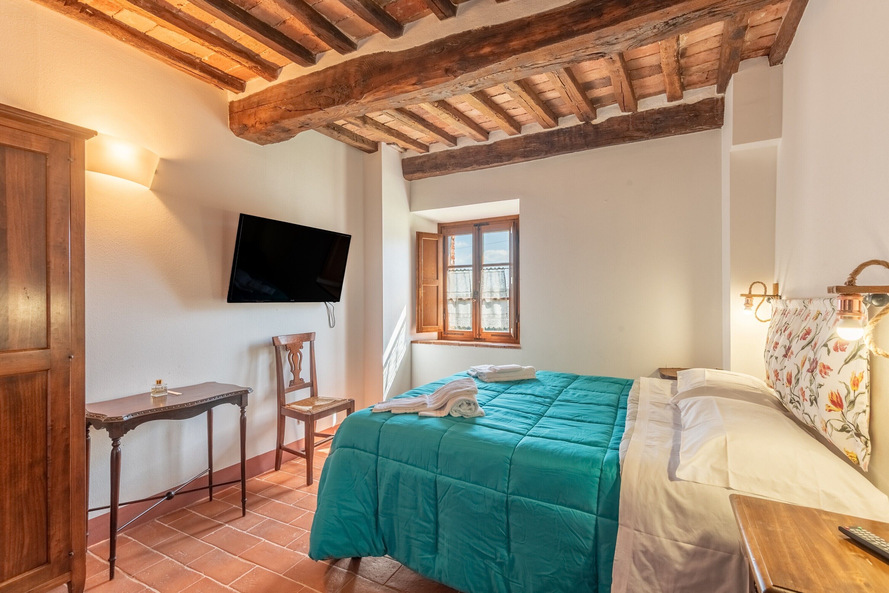 Un dormitorio con techo de madera, una cama con colcha verde, un televisor montado en la pared y una ventana con cortinas.