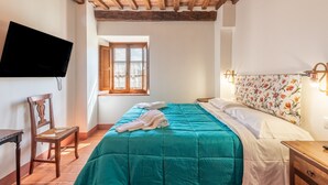 A bedroom with a wooden ceiling, a bed with a teal bedspread, a chair, a TV mounted on the wall, and a window with curtains. - Room 'Pernina' – Agriturismo la Taverna di Celsa with Shared Pool (Sovicille)