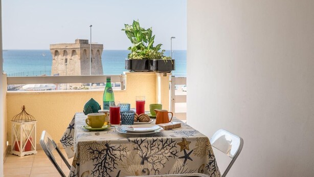 A balcony with a table set for breakfast, overlooking a coastal view with a historic tower in the background. - Desire for the Sea (Gallipoli)