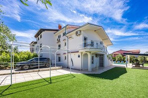 Ein zweistöckiges Haus mit Balkon, ein Auto auf einem überdachten Parkplatz und ein Schwimmbad im Hintergrund.
