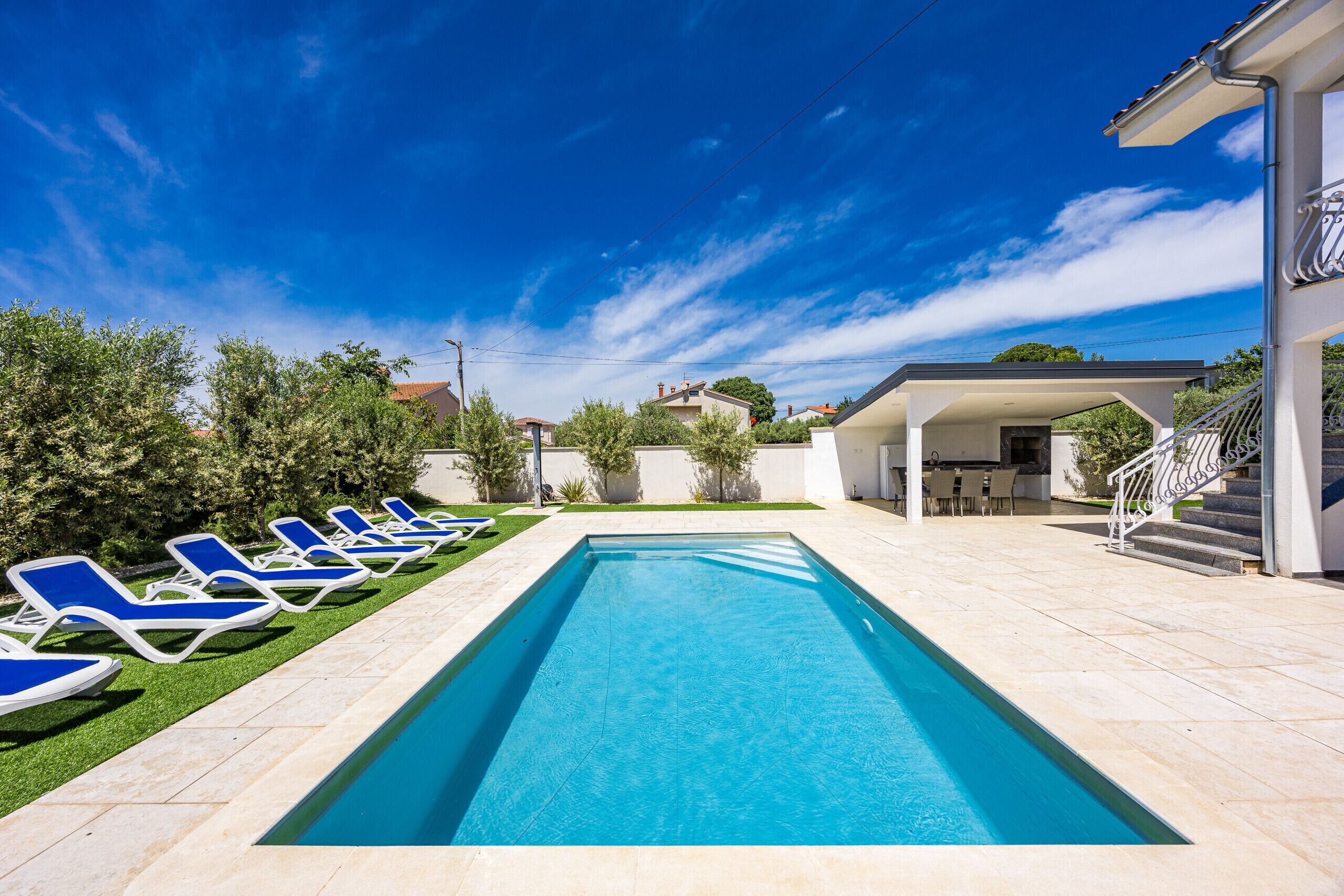 A rectangular swimming pool with blue water, surrounded by a tiled patio. There are blue lounge chairs aligned along the pool's edge.