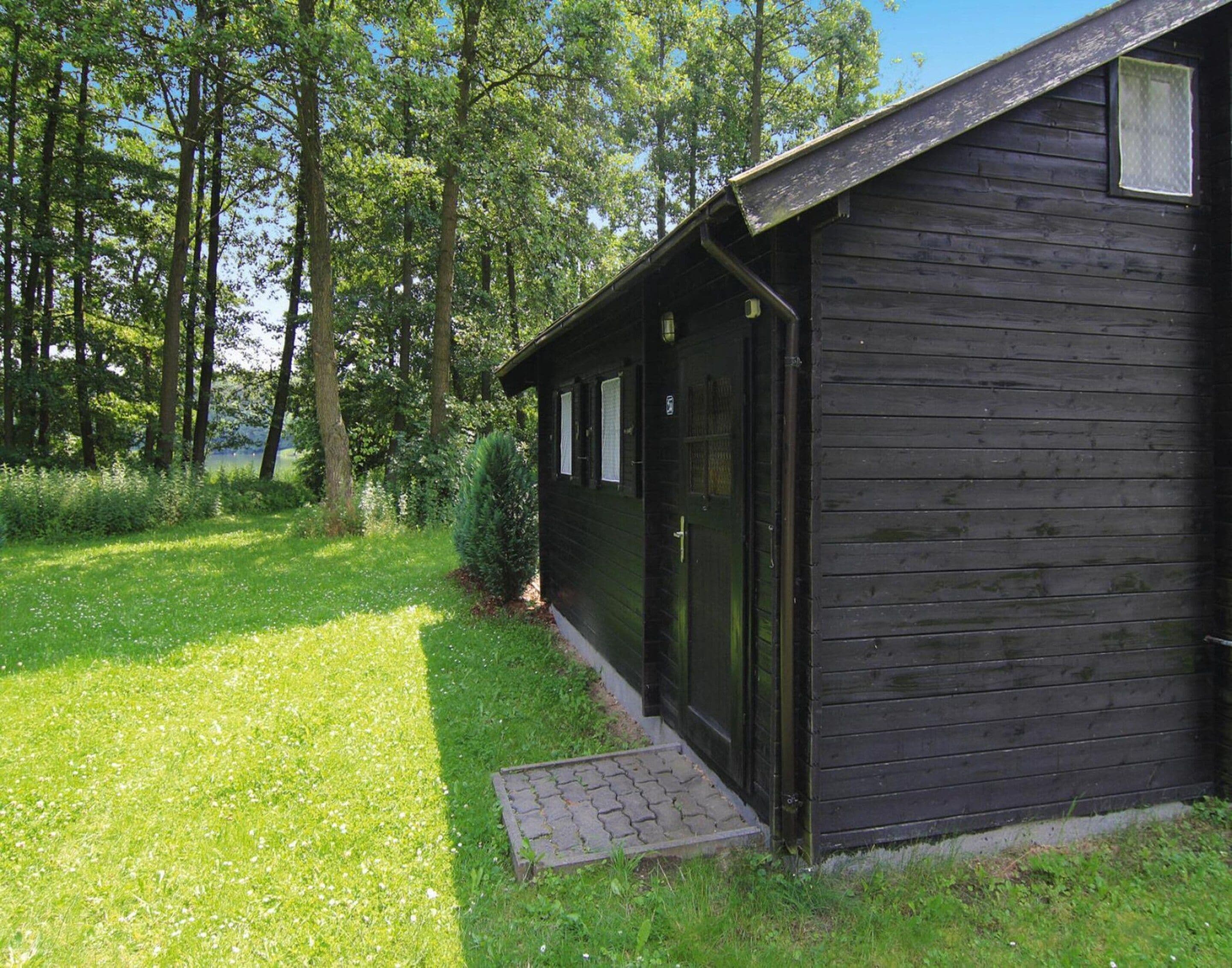 A small, dark wooden cabin with a single door and windows, surrounded by a grassy area and trees.