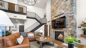 Amplia sala de estar con chimenea de piedra, sofá de cuero y escalera con barandilla negra.