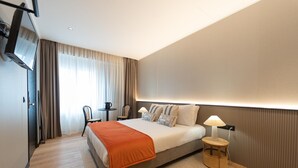 Una habitación de hotel moderna con una cama grande, un televisor montado en la pared, una mesita con una lámpara y una silla.