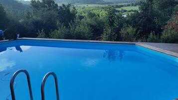 A clear blue swimming pool with metal ladders, surrounded by lush greenery and hills in the background.