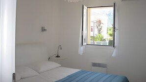 A bedroom with a bed, a bedside table with a lamp, a window with a view, and a ceiling light fixture. - Modern T3 Apartment in Calvi with Terrace – Available from August 22, Discounted Rates (Calvi)