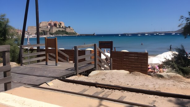 A beach with a wooden walkway leading to the water, a sign reading 'MARINEPORT,' and a distant view of a historic building. - Modern T3 Apartment in Calvi with Terrace – Available from August 22, Discounted Rates (Calvi)