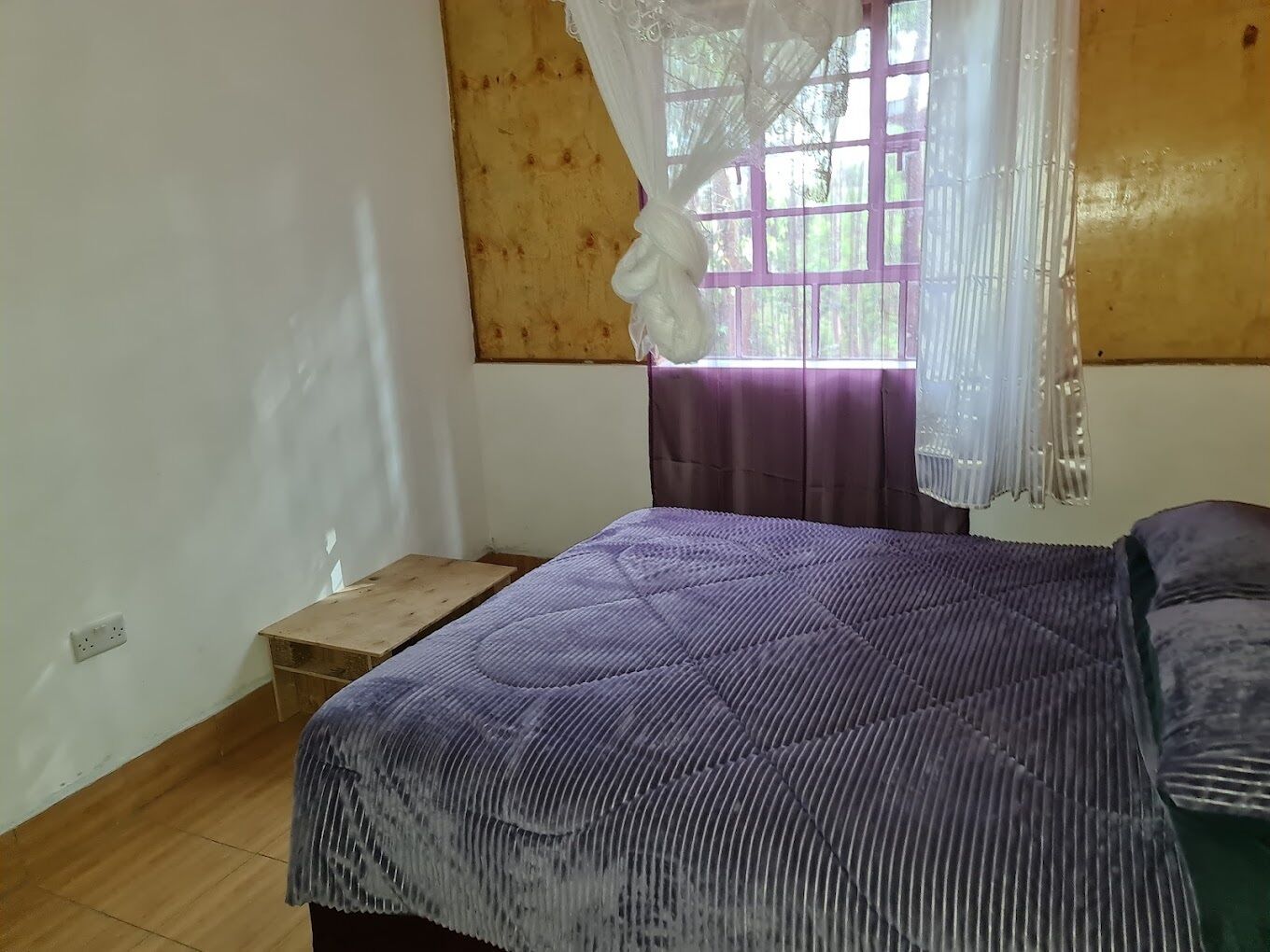 A bedroom with a wooden floor, a bed with a purple quilt, a window with purple curtains, and a wooden headboard.