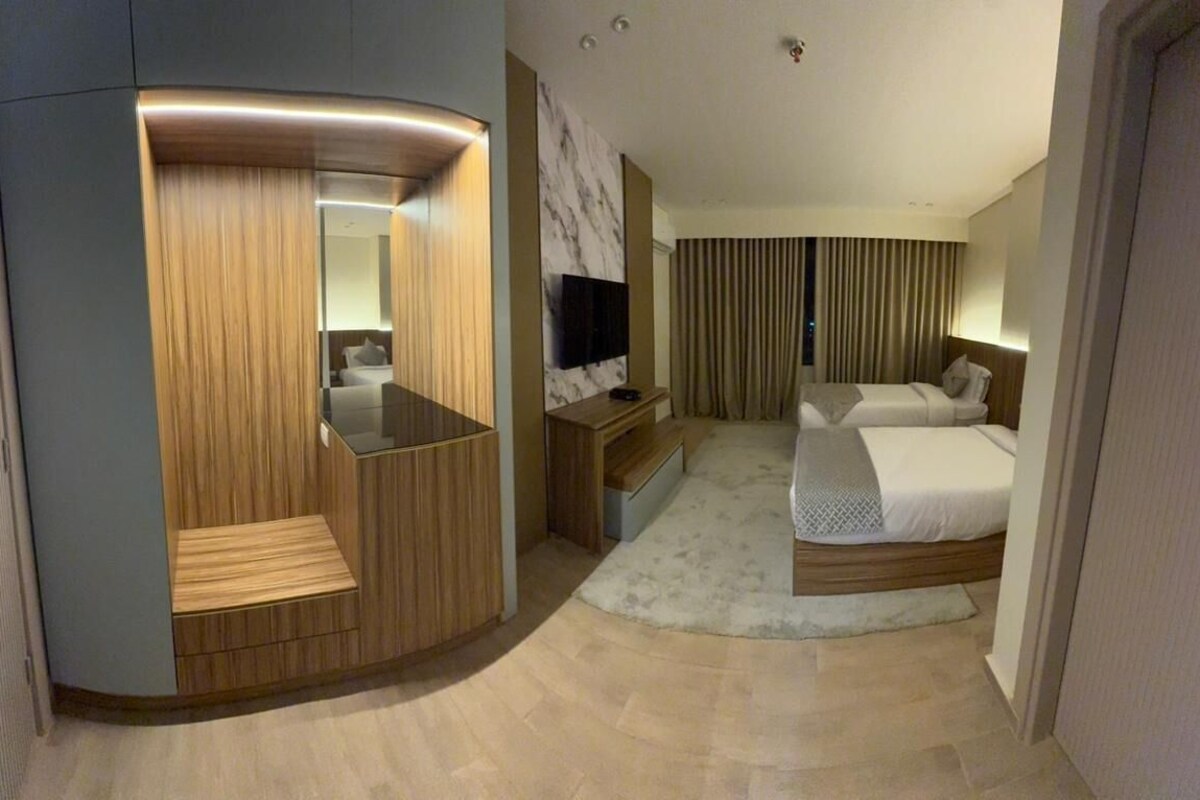 Una habitación de hotel moderna con una zona de estar curva, un televisor de pantalla plana, una cama con colcha blanca y un ventanal con cortinas.