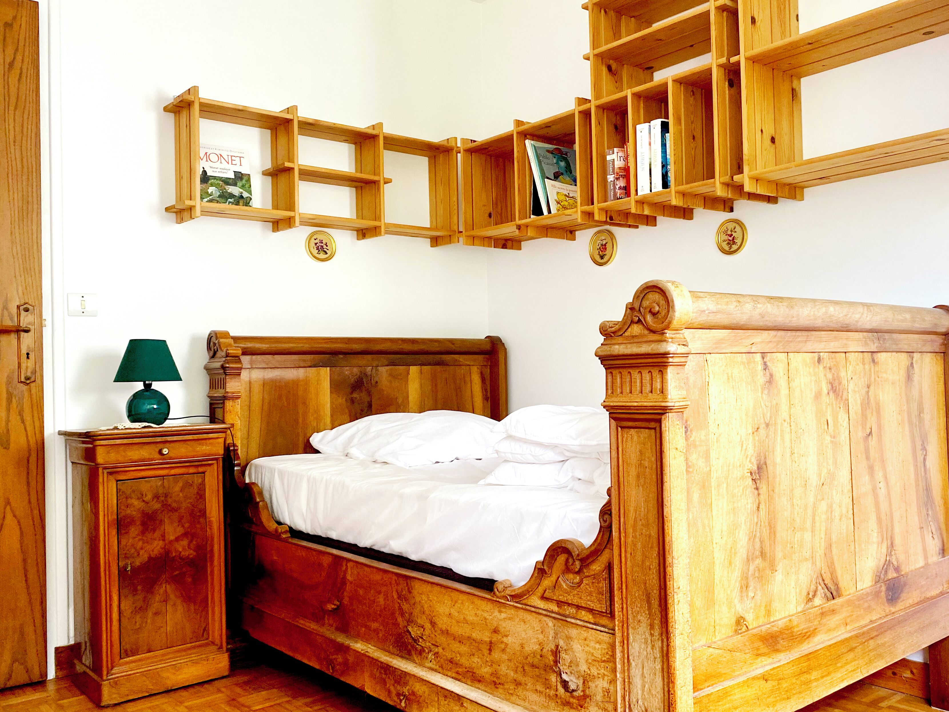 A wooden bed with a headboard, a bedside table with a lamp, and a wooden shelf above the bed.