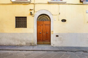 Um edifício amarelo com uma porta dupla de madeira, uma pequena janela com grade de metal e um pavimento de azulejos.