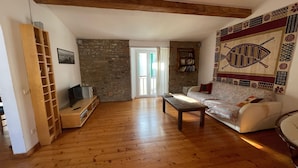 Ein Wohnzimmer mit Holzboden, einer Couch, einem Couchtisch, einem Fernseher auf einem Holzboden und einer Steinwand, sowie einem gemusterten Wandteppich.