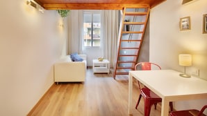 Ein Zimmer mit Holzboden, einem weißen Sofa, einem kleinen Tisch mit einer Obstschale, einem roten Sessel, einem weißen Tisch und einer an der Wand stehenden Leiter.