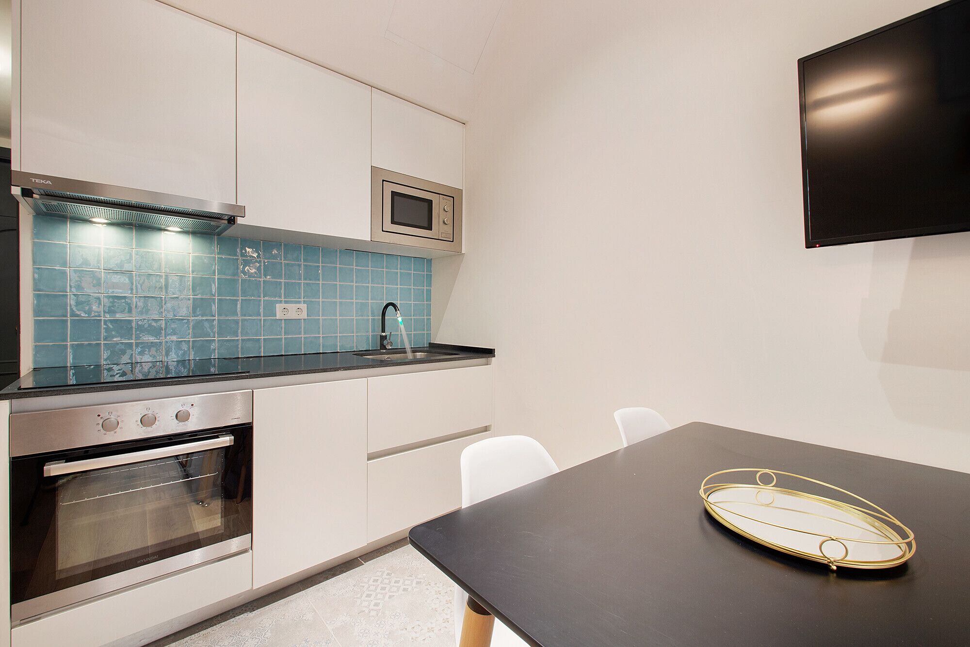 Een moderne keuken met een magnetron, oven en een blauwe betegelde spatwand. Er is een zwart aanrecht en een tafel met een gouden dienblad.