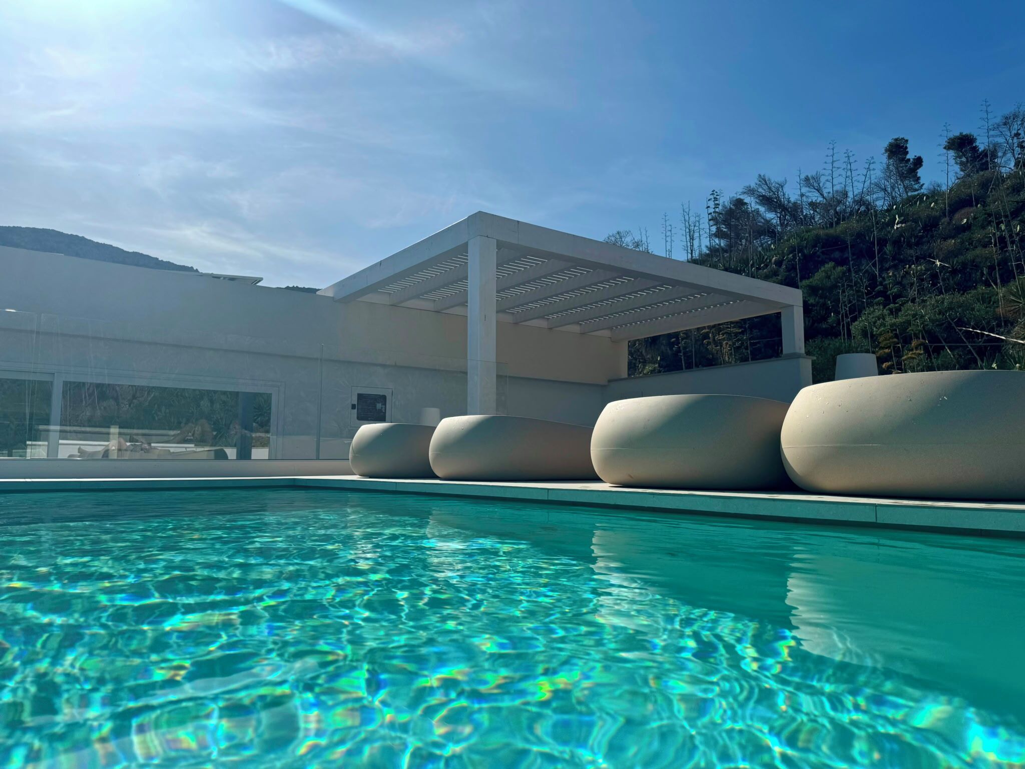 Una piscina con agua cristalina, un edificio moderno de fachada blanca y un área exterior cubierta con asientos.
