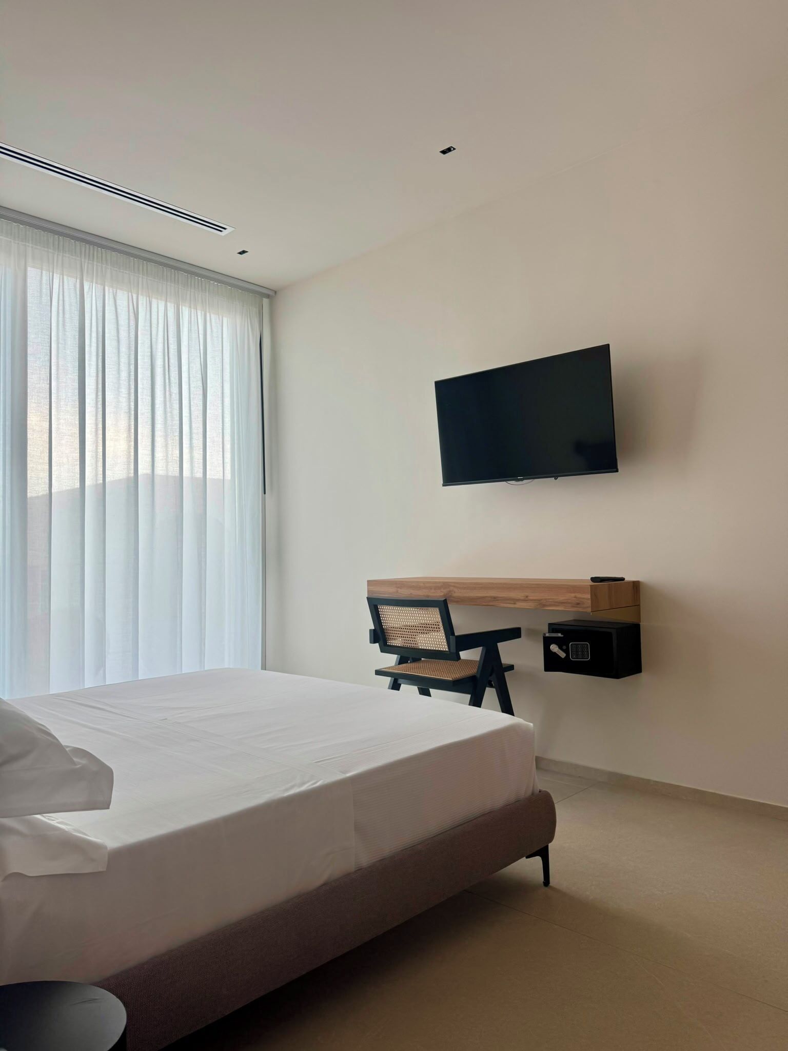 A hotel room with a bed, a TV mounted on the wall, a desk with a chair, and a safe.