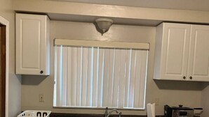 A kitchen with white cabinets, a window with blinds, a sink, and a ceiling light fixture.