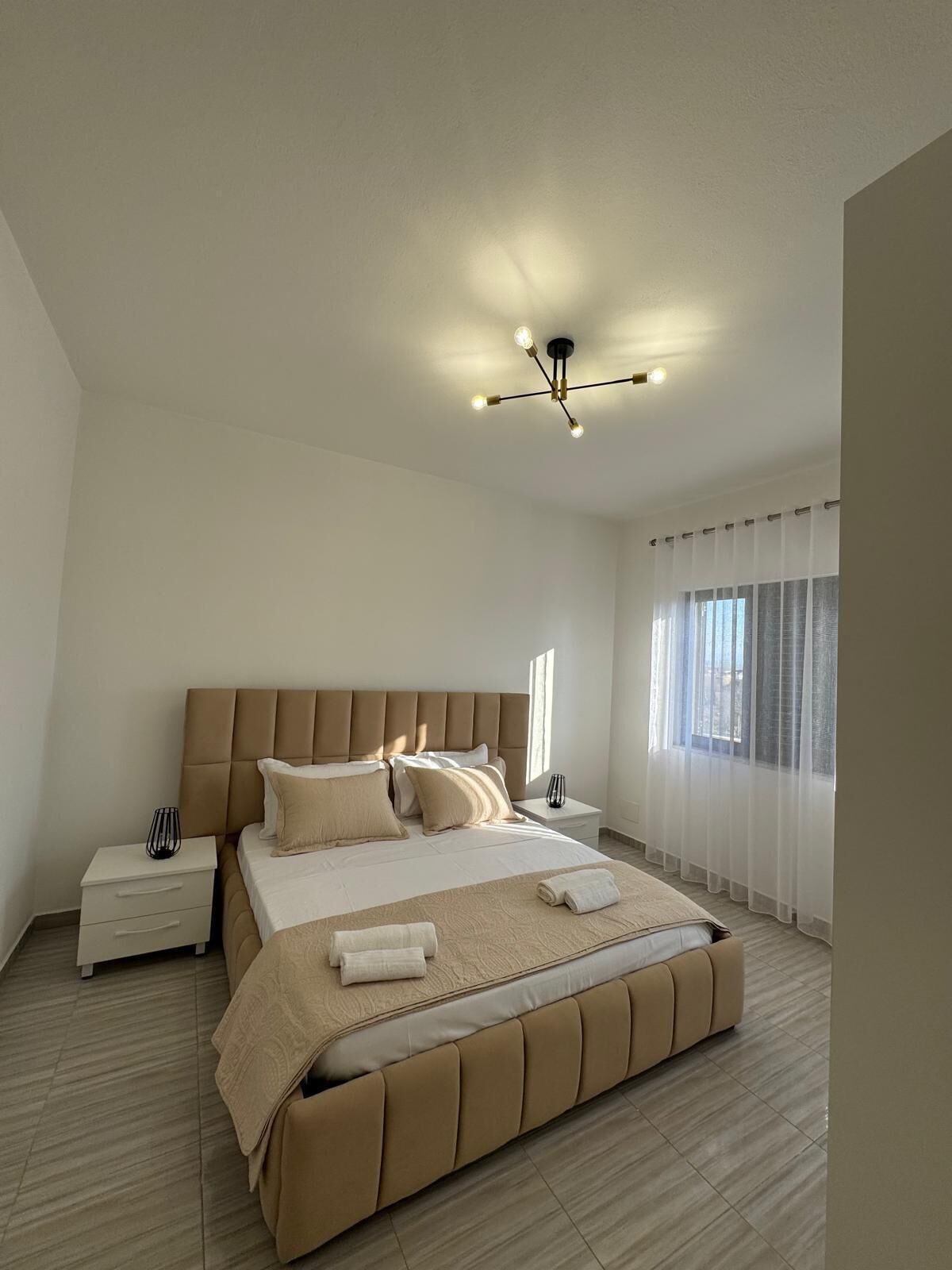 Une chambre moderne avec un grand lit, une table de chevet, une commode, un luminaire au plafond et une fenêtre avec des rideaux.