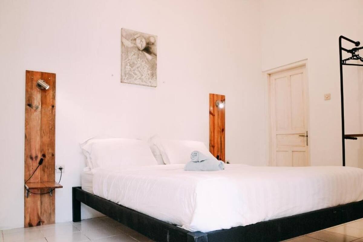 A single-bed room with a wooden headboard, a bedside table, a wall-mounted artwork, and a white door.