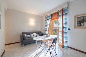 A modern living room with a grey sofa, a round white table, and two chairs. There is a window with blue curtains and a view of the outside. - Sanremo la Mimosa by Wonderful Italy (Sanremo)