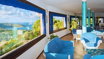 Un balcón con vistas a un paisaje costero, sillas tapizadas en azul y una mesa con lámpara.