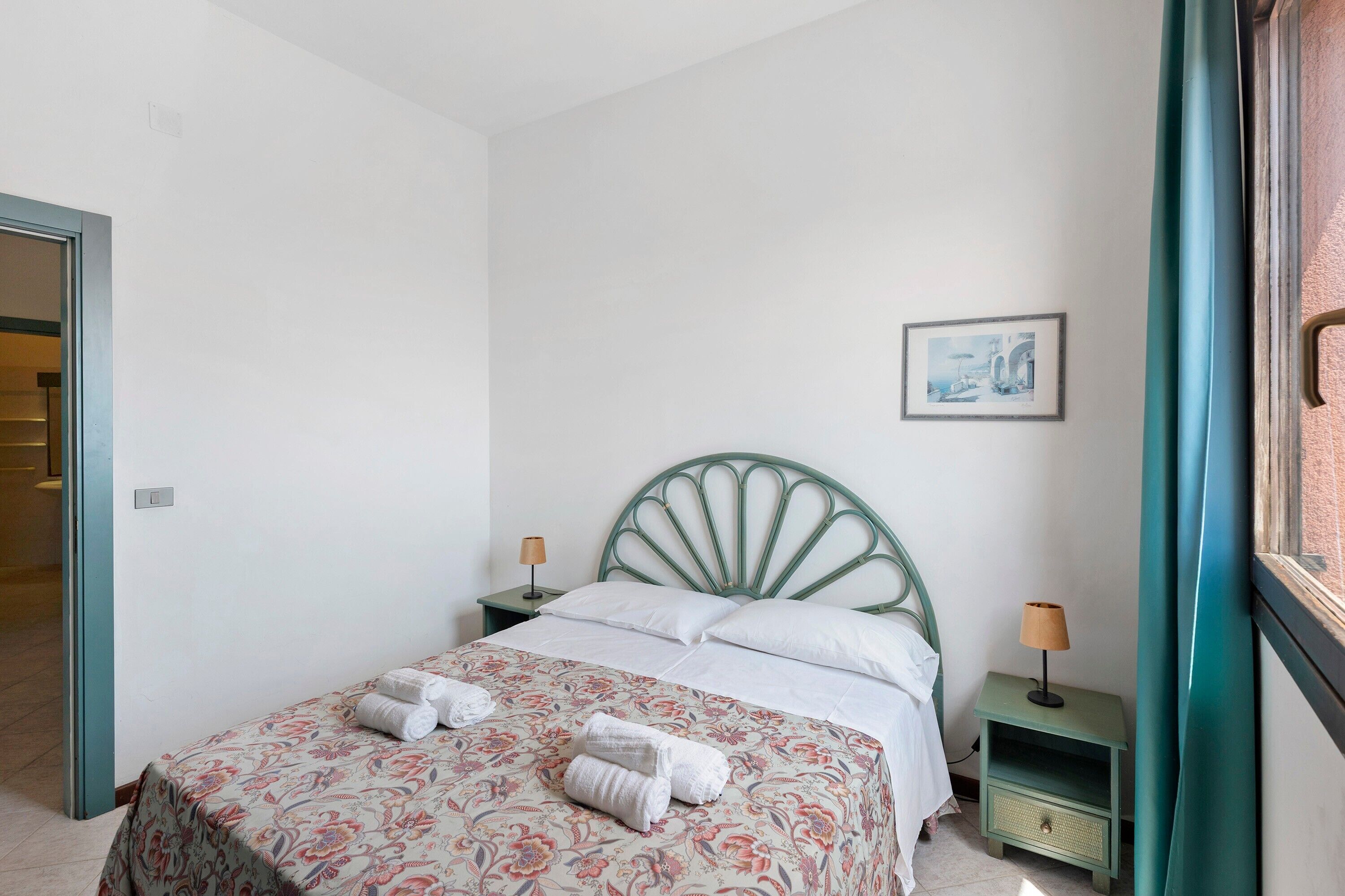 Un lit bien fait, recouvert d’une couvre-lit à motifs floraux, avec deux serviettes enroulées, une tête de lit en métal vert, une petite table de chevet avec une lampe et un tableau encadré accroché au mur.