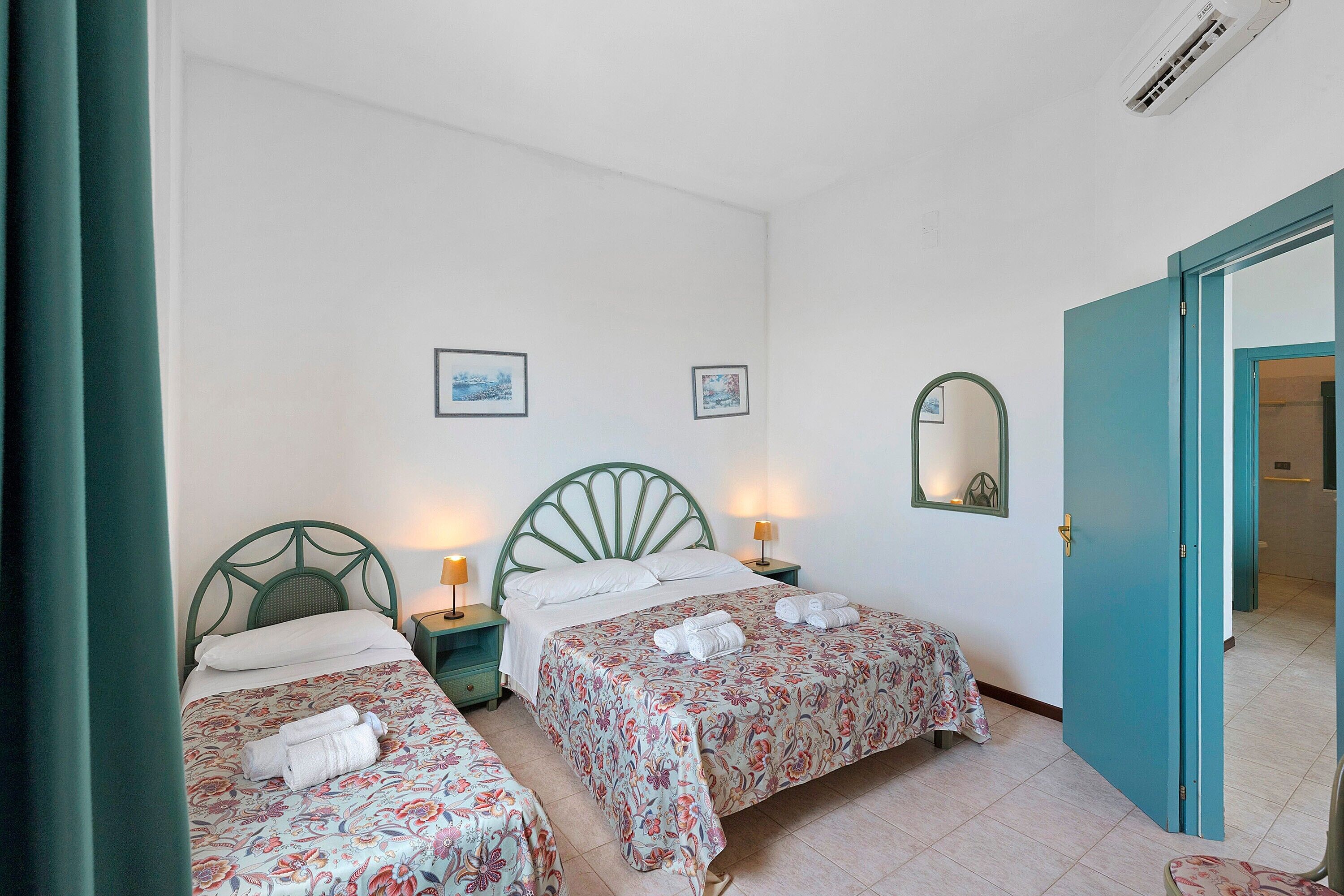 A room with two beds, each with floral bedding and towels, a green headboard, bedside lamps, and a mirror on the wall.