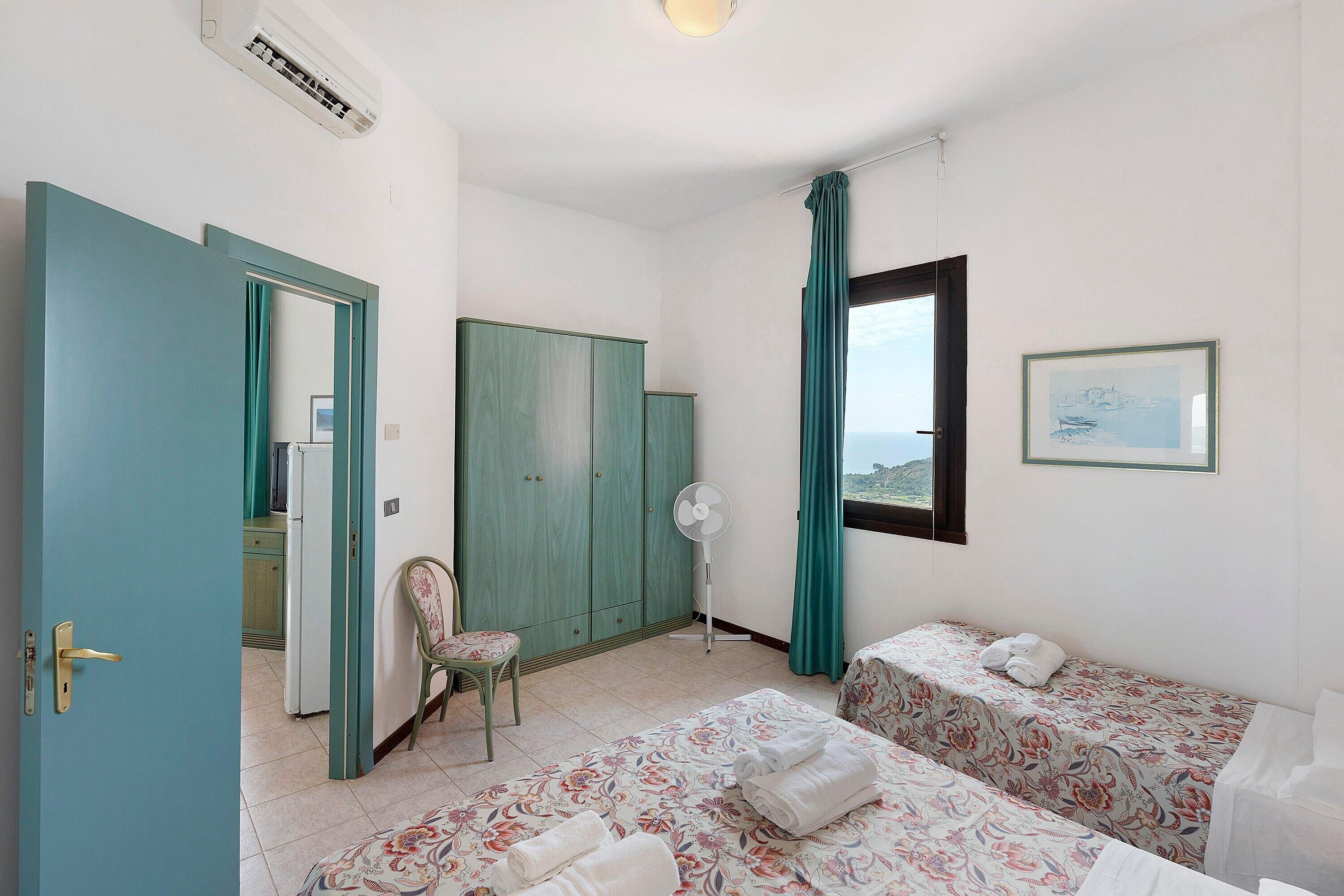 A room with two beds, a chair, a fan, and a window with curtains.