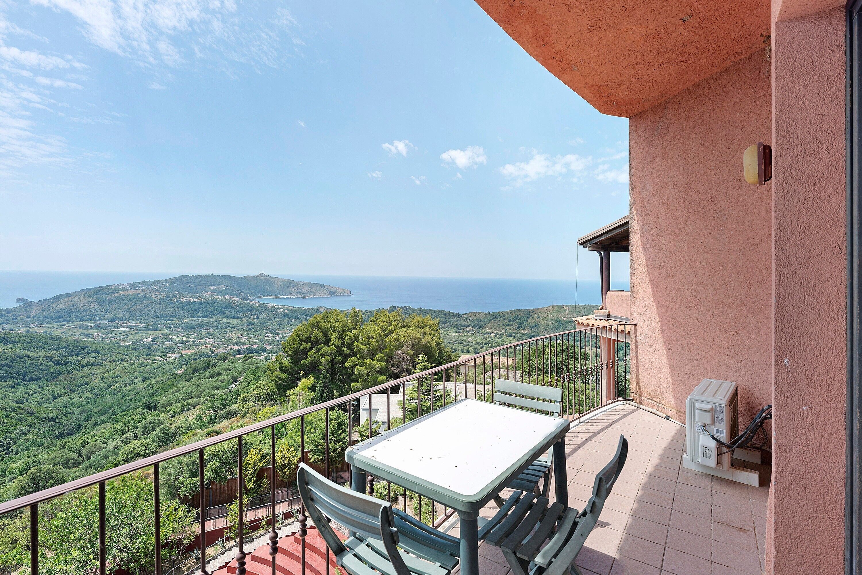 Un balcon avec une table et des chaises, donnant sur un paysage pittoresque composé de collines et d’une étendue d’eau.