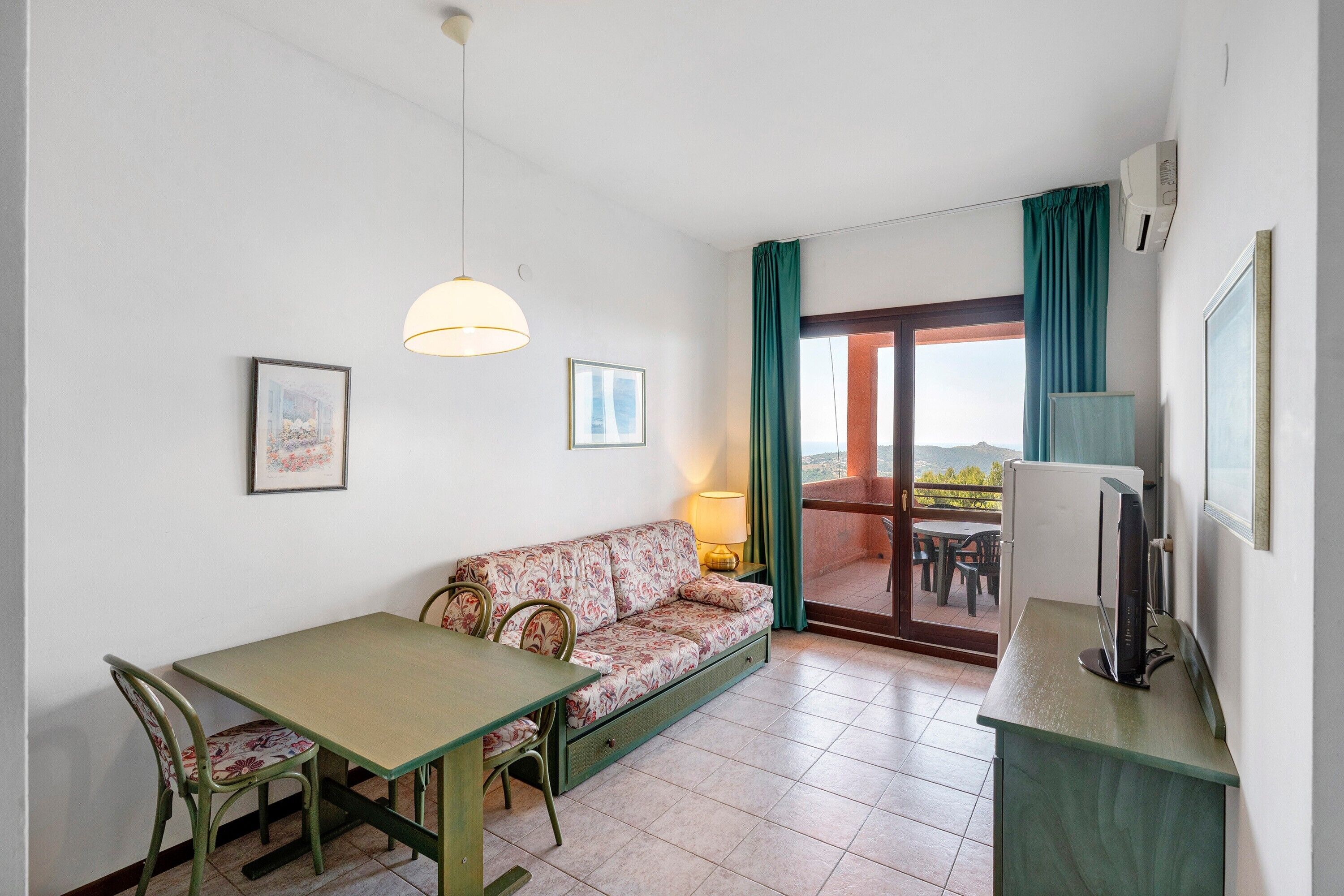Een kamer met een bank, eettafel, stoelen, een televisie en een balkon met gordijnen.