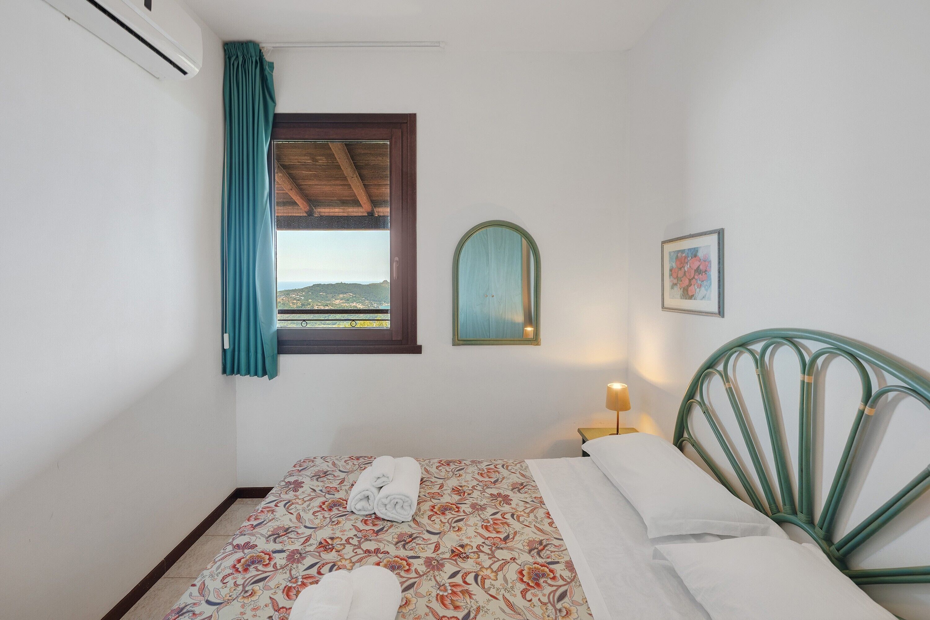 Een slaapkamer met een bed, een raam met gordijnen, een aan de muur hangend schilderij en een nachtkastje met een lamp.
