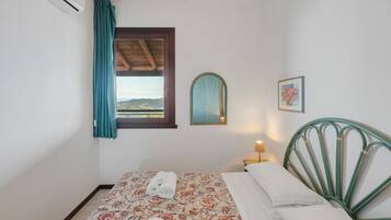 Een slaapkamer met een bed, een raam met gordijnen, een aan de muur hangend schilderij en een nachtkastje met een lamp.