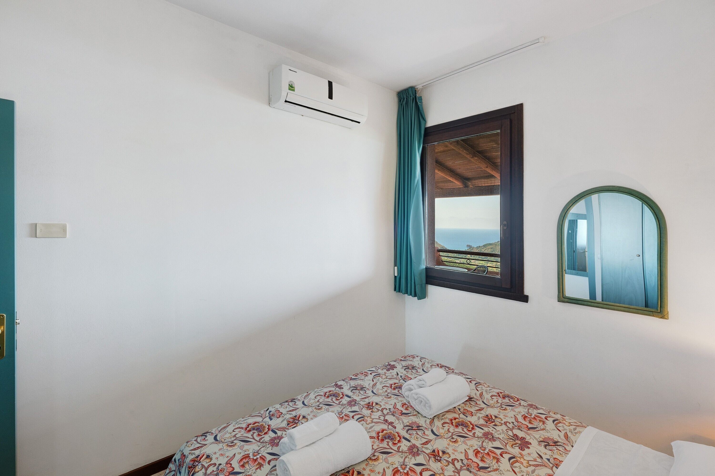 Un lit simple avec une literie à motifs floraux, un appareil de climatisation, une fenêtre avec vue et un miroir au mur.