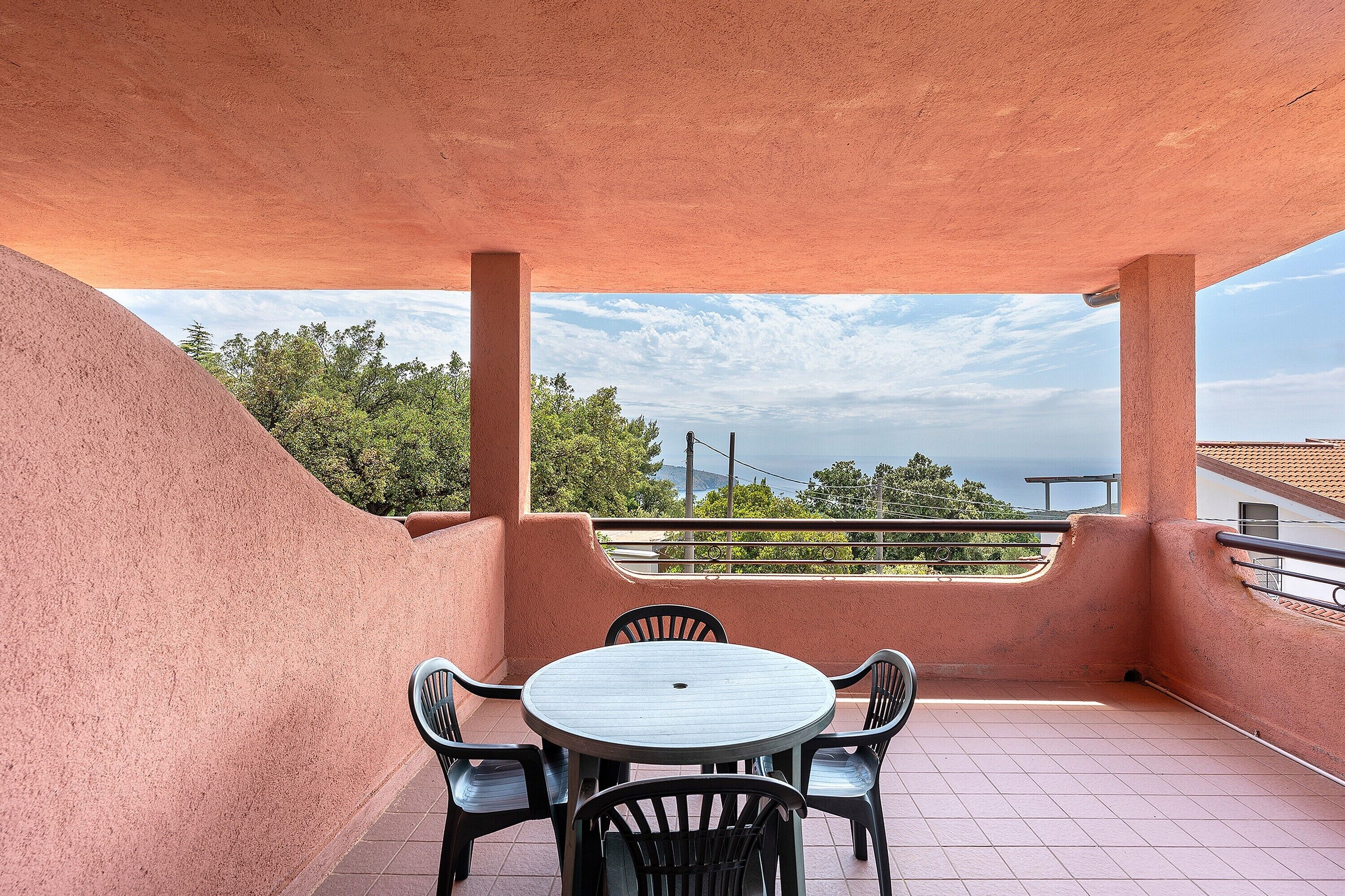 A balcony with a round table and chairs, a view of trees and a distant landscape, and a red textured wall.