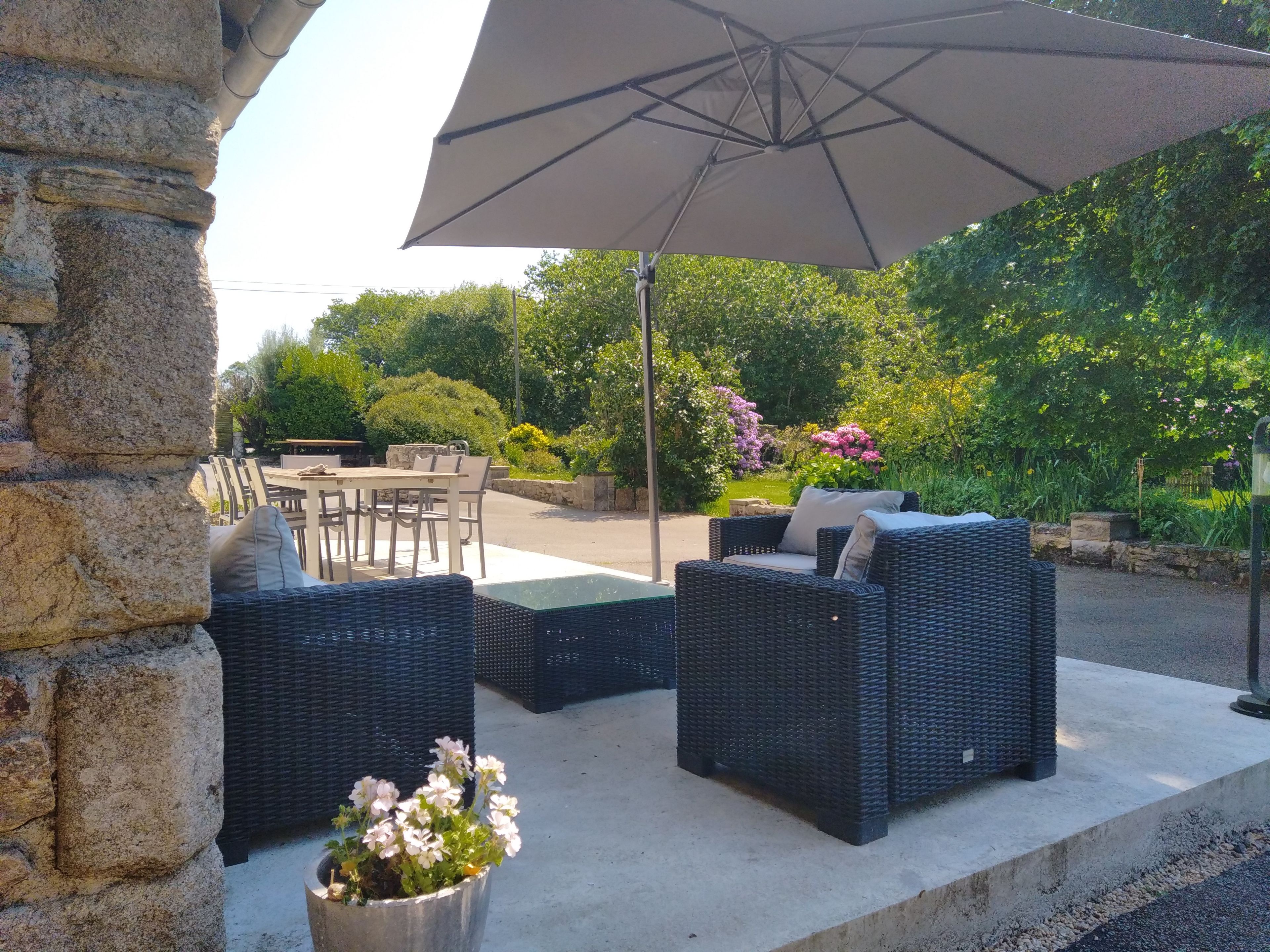 An outdoor seating area with wicker furniture and a large umbrella, surrounded by greenery and flowers.