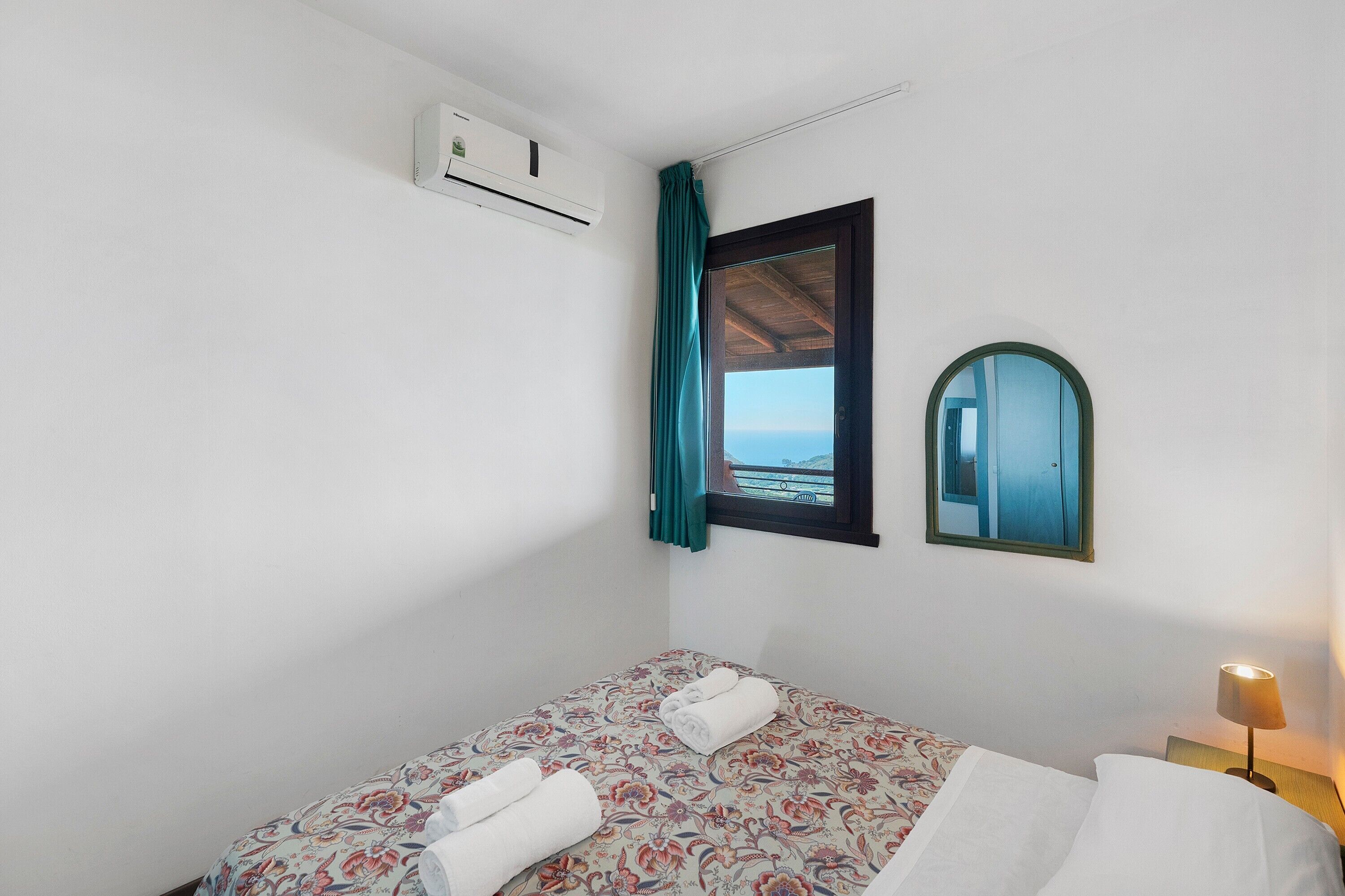 Un dormitorio con cama, ventana con cortinas, espejo, lámpara de noche y aire acondicionado.