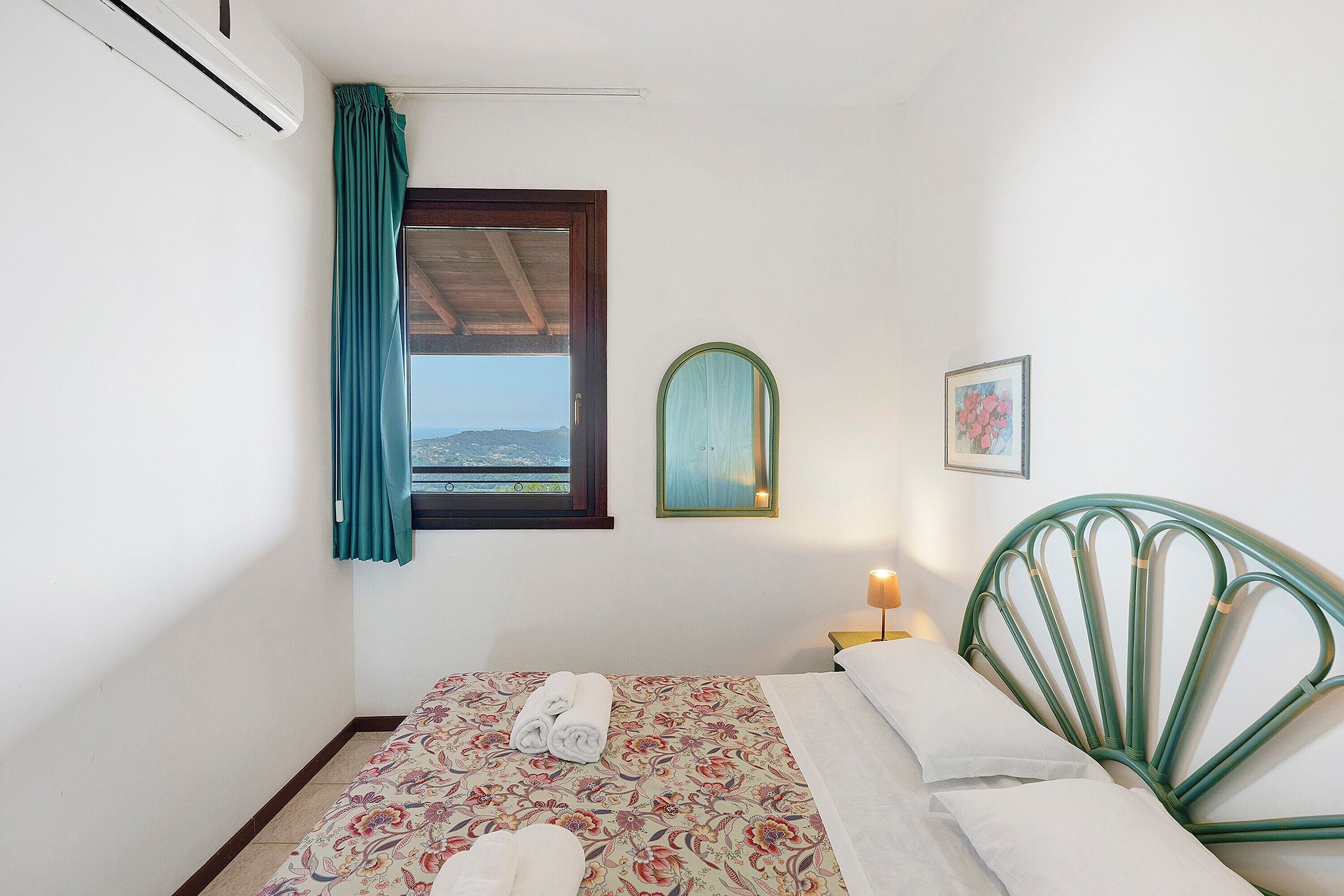 A bedroom with a bed, a window with curtains, a bedside lamp, and a framed picture on the wall.