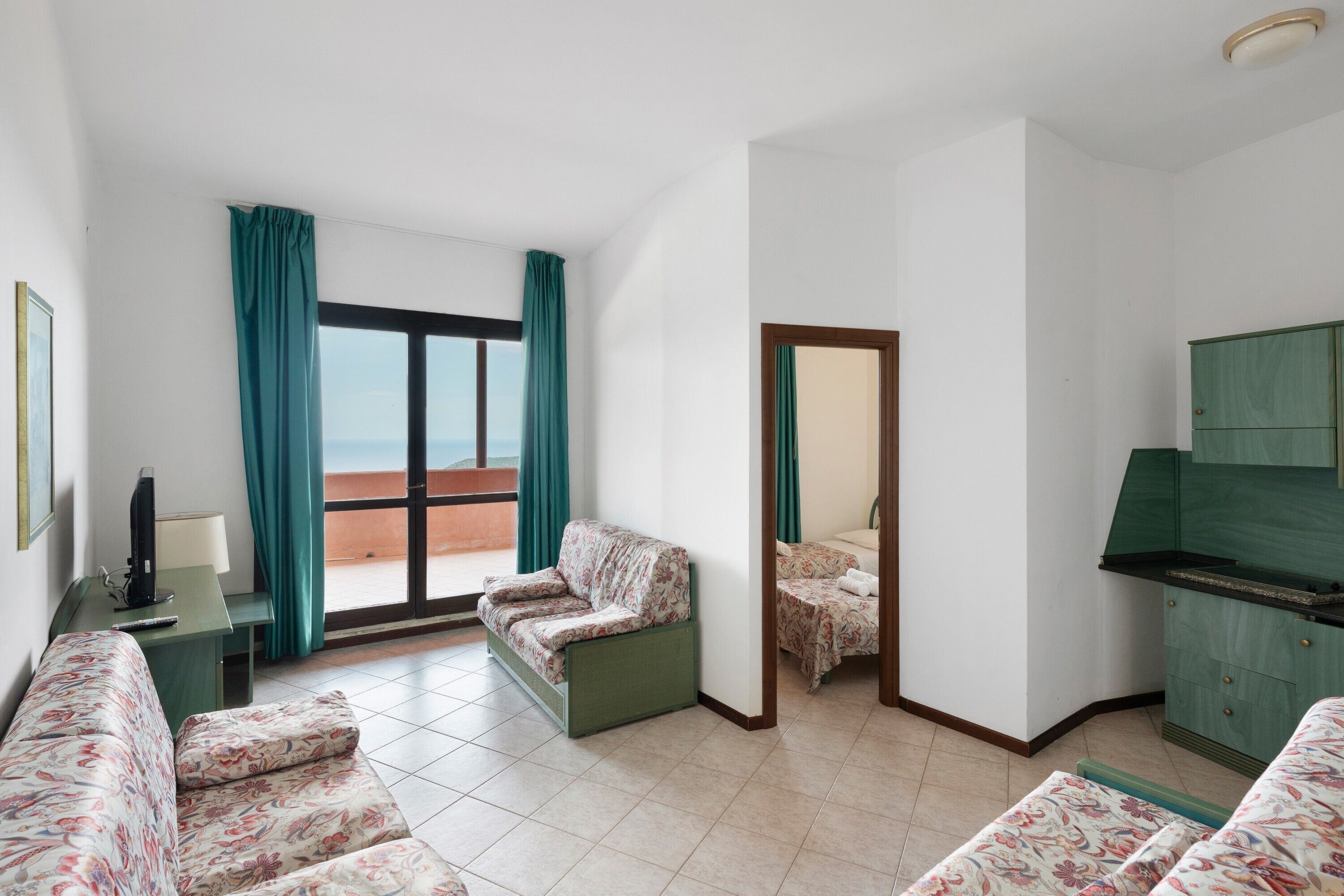 Une chambre d’hôtel avec un canapé, une petite table, une télévision, un balcon avec des rideaux et une armoire verte.