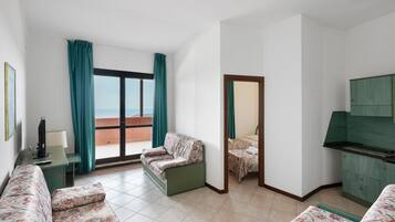Une chambre d’hôtel avec un canapé, une petite table, une télévision, un balcon avec des rideaux et une armoire verte.