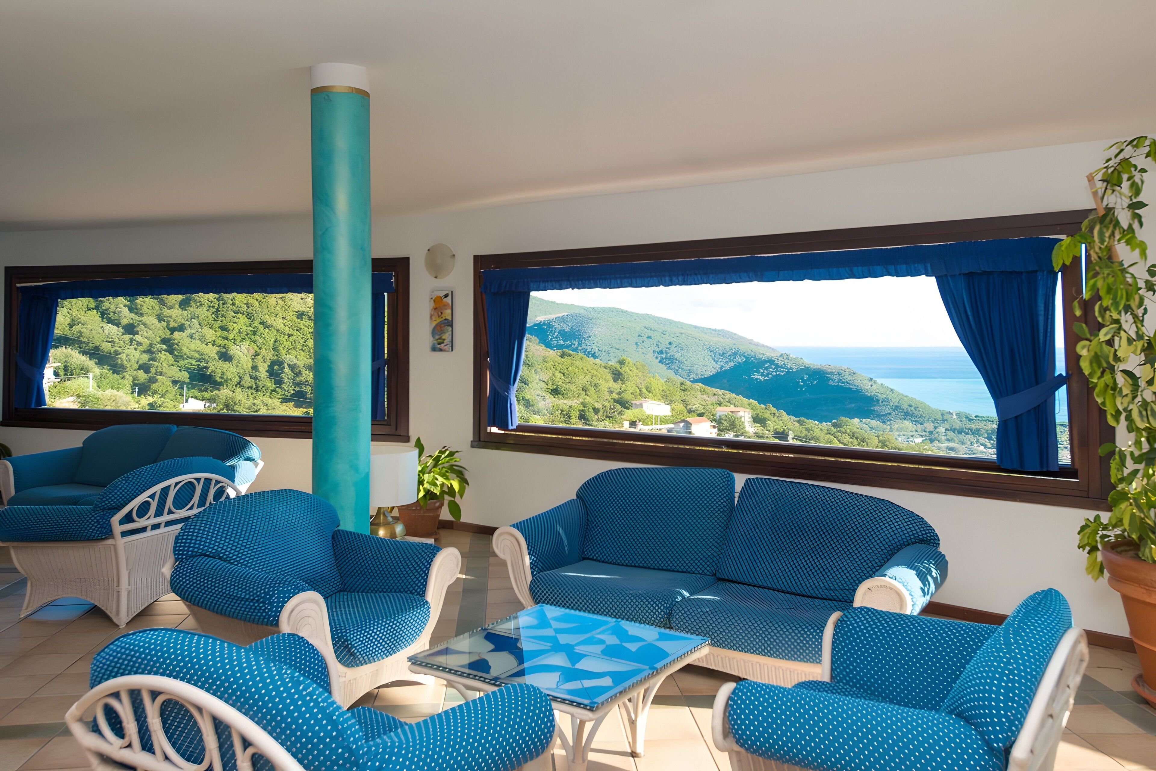 A room with a view of green hills and the sea, furnished with blue and white furniture, and a large window with curtains.
