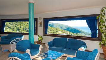 A room with a view of green hills and the sea, furnished with blue and white furniture, and a large window with curtains.